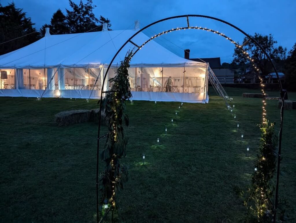 A white marquee in a field with fairy lights around an archway. It's dusk.