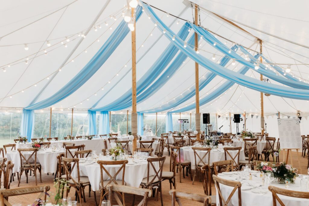 Inside a wedding marquee with wooden chairs and white tablecloths. Bunting and blue drapes hang from the ceiling.